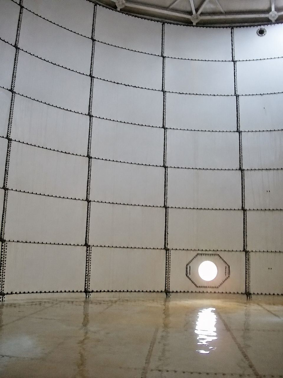 Interior of an empty bolted-steel storage tank showing panel seams and a manway
